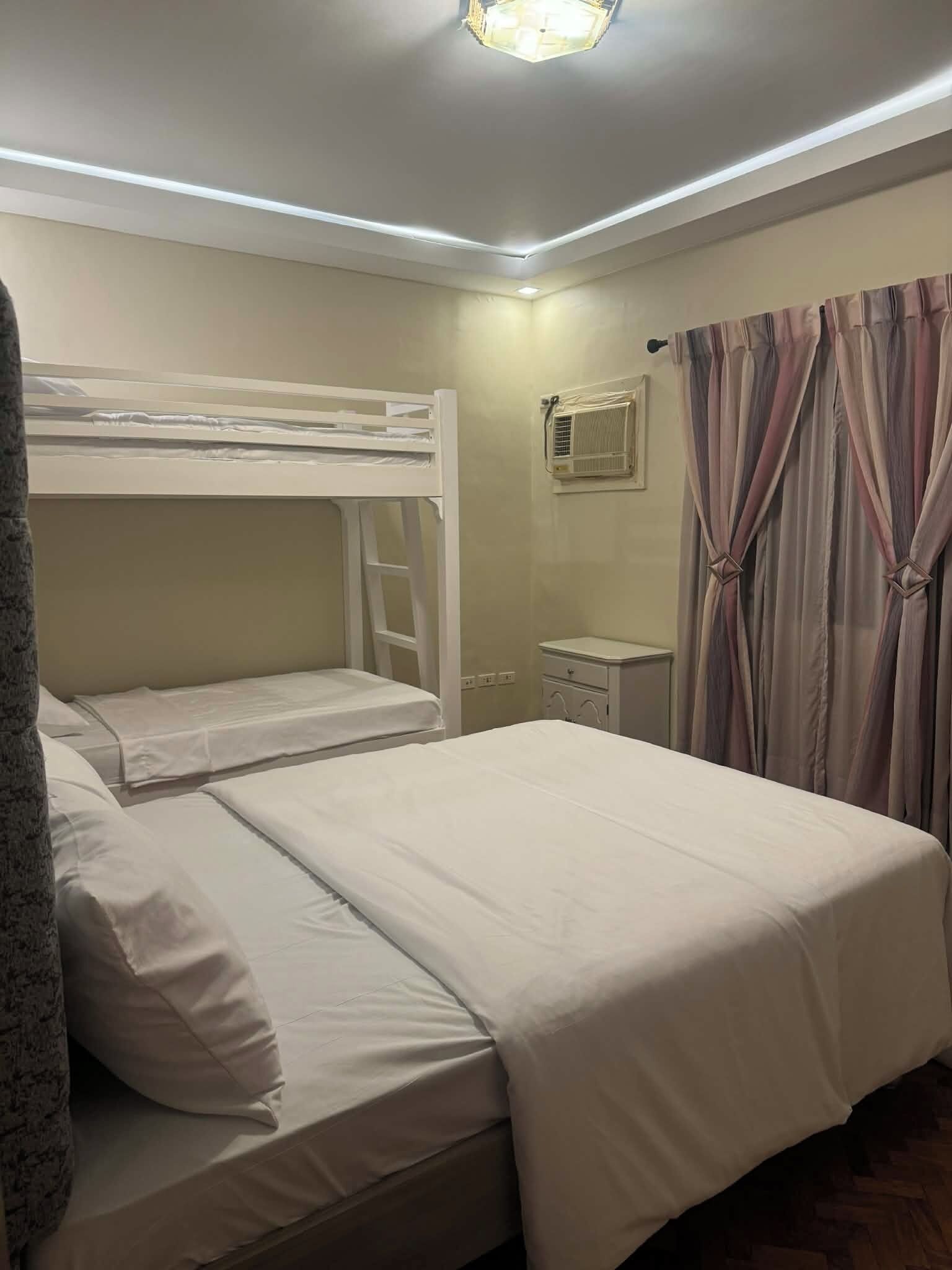 A bunk bed room with a white bed, a small bedside table, an air conditioning unit, and a curtain with a knot detail.