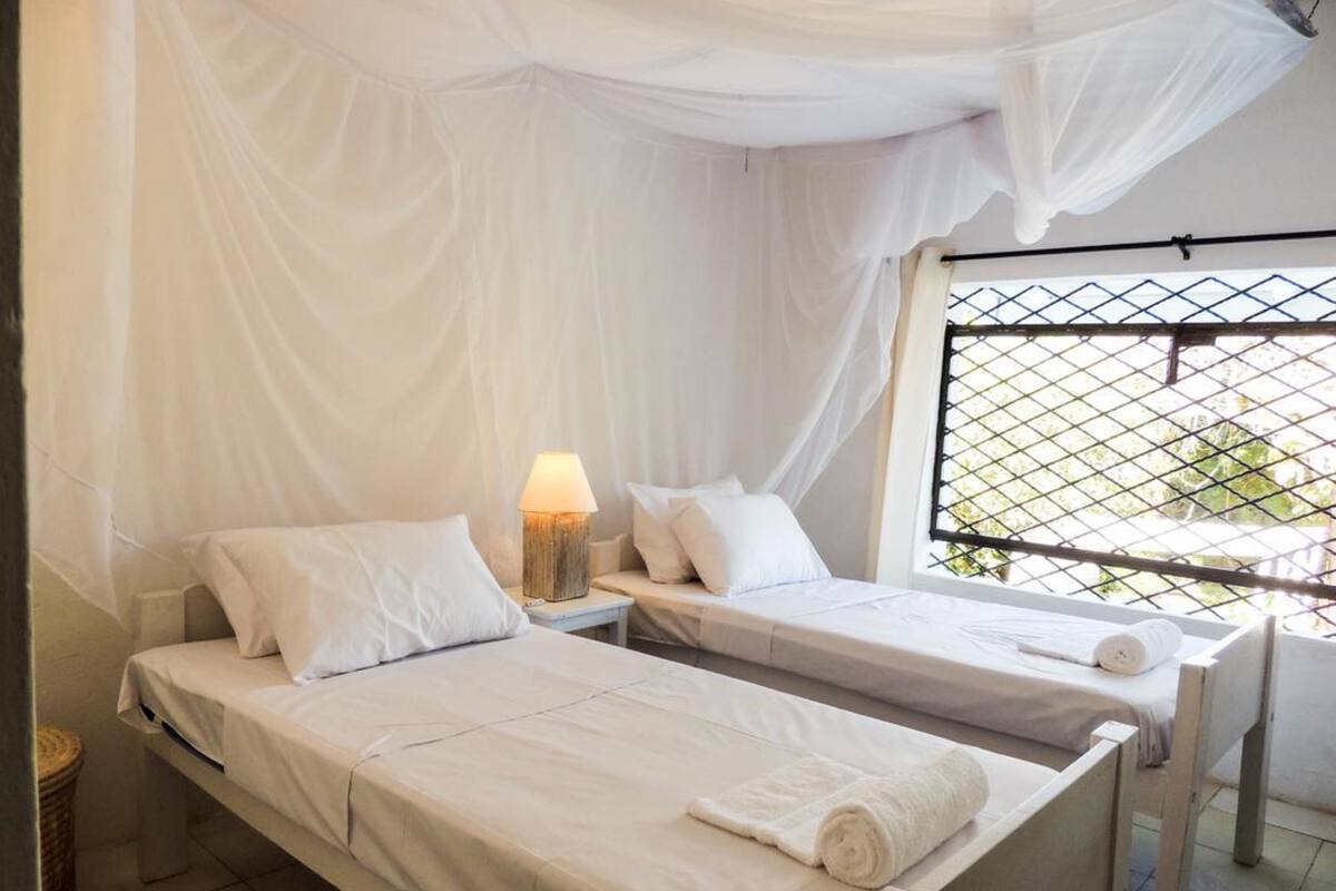 Habitación con dos camas, una mesita de noche con lámpara, una ventana con reja de metal y un dosel blanco sobre las camas.