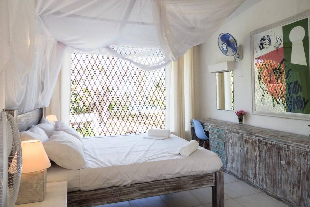 Un dormitorio con cama, una mesita de noche con lámpara, cómoda, ventana con vistas y un cuadro en la pared.