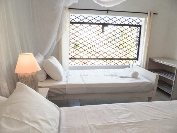 Un dormitorio con cama, lámpara de noche, ventana con mosquitero y cortina blanca.