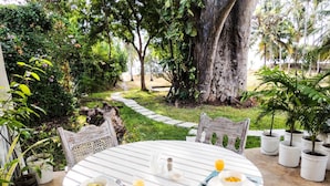 Una mesa redonda blanca con un desayuno servido, rodeada de sillas, en un jardín exuberante con un sendero de madera y diversas plantas.