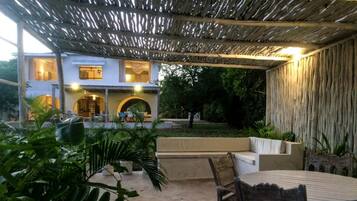 Un patio al aire libre con una mesa y sillas, rodeado de plantas en macetas y un espacio de descanso.