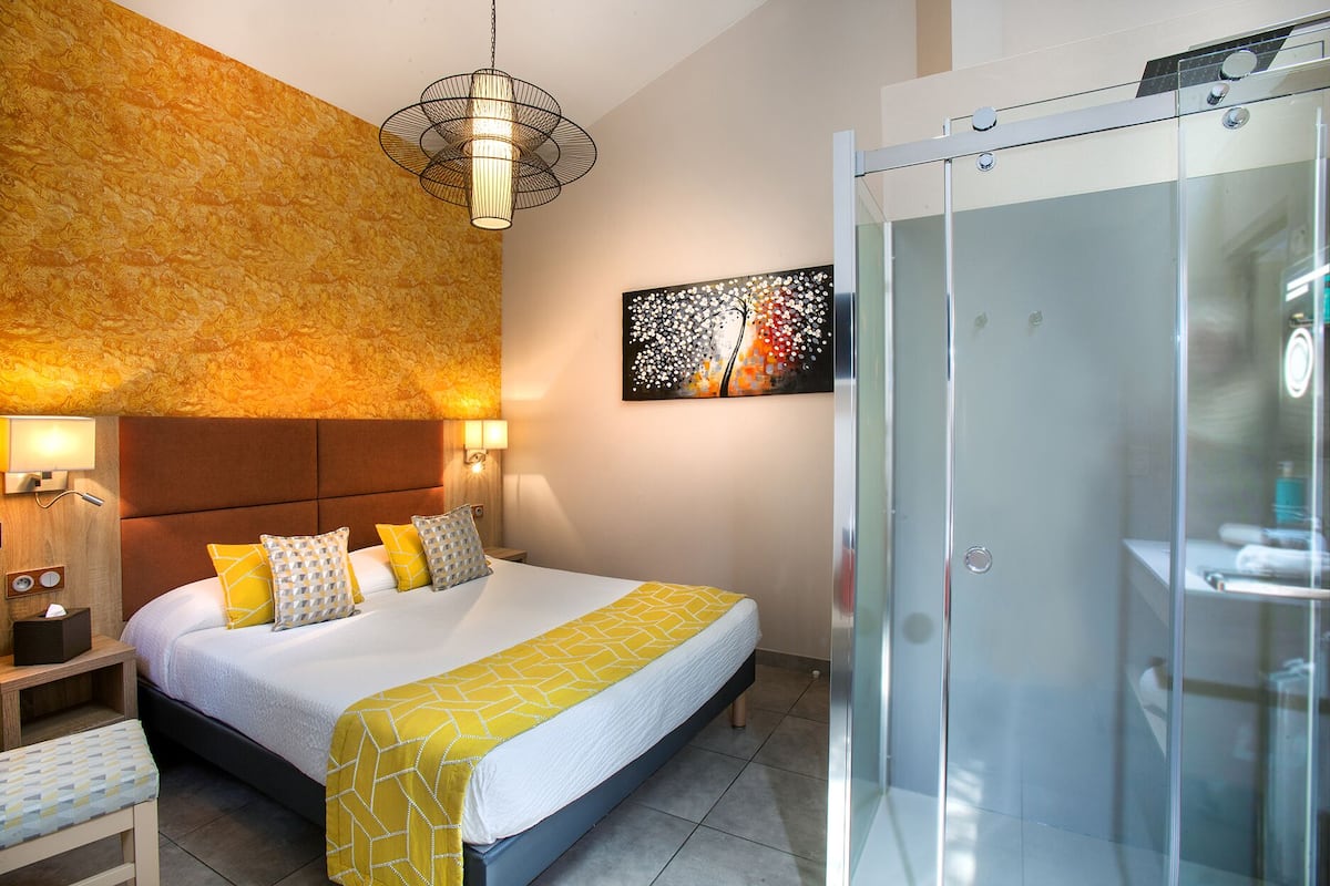 A hotel room with a bed, bedside table, lamp, and a glass shower enclosure.