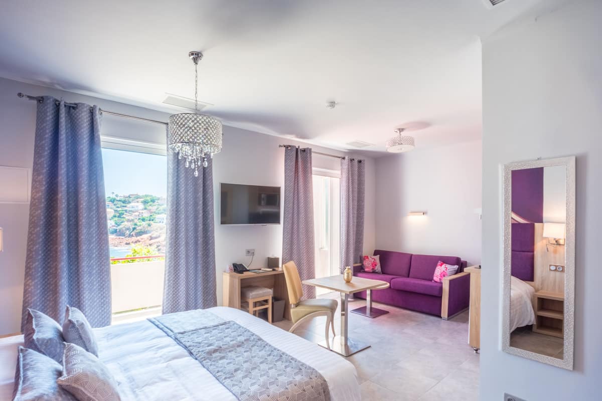 A modern hotel room with a large bed, a desk with a chair, a purple sofa, and a TV mounted on the wall.