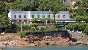 A hotel with a restaurant named "Les Flots Bleus" on the second floor, situated on a hillside with a rocky beach in the foreground. - Les Flots Bleus (Saint-Raphael)