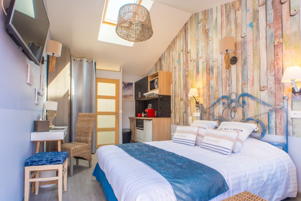 A bedroom with a bed, a desk, a chair, a TV, and a kitchenette.