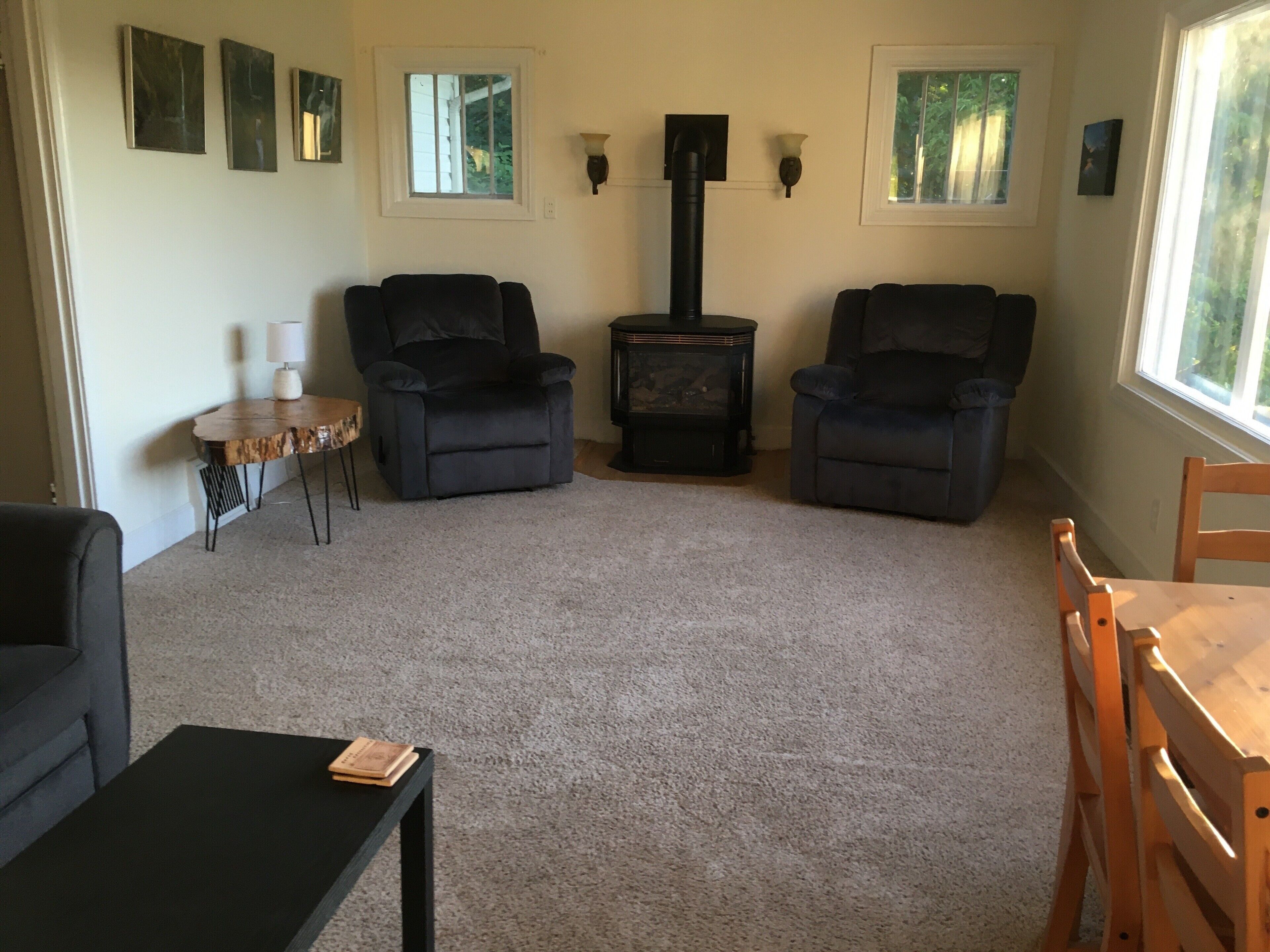 A living room with two black armchairs, a small round table with a lamp, a wood-burning stove, and a window with a view of greenery.