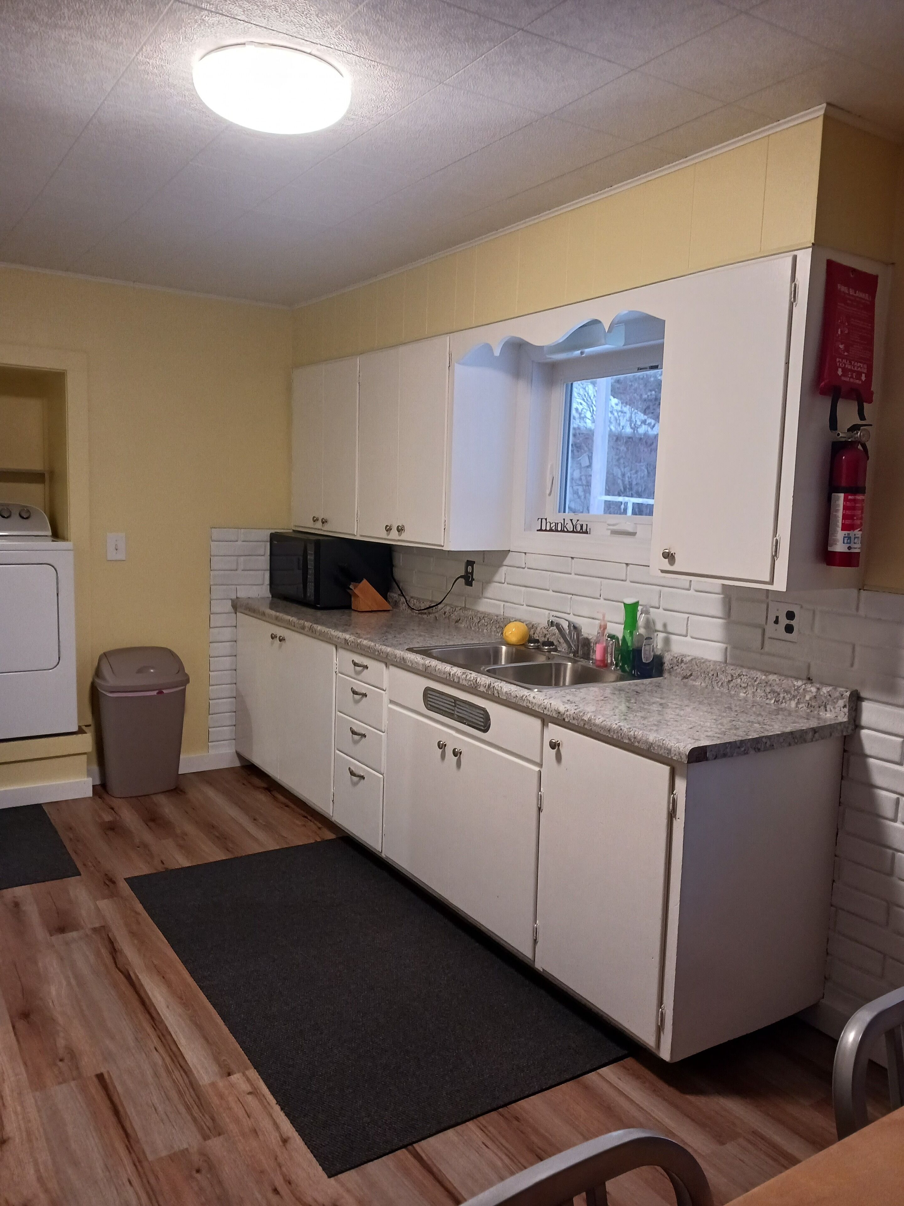 Een kleine keuken met witte kastjes, een magnetron en een spoelbak. Er is een raam met een bord waarop 'Thank You' staat, een brandblusser en een prullenbak.
