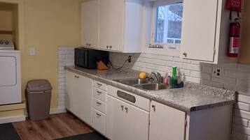 Een kleine keuken met witte kastjes, een magnetron en een spoelbak. Er is een raam met een bord waarop 'Thank You' staat, een brandblusser en een prullenbak.