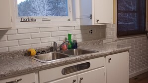 Una cocina con armarios blancos, encimera de granito, ventana con un cartel que dice 'Gracias' y vistas a árboles fuera.