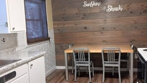 Una cocina con gabinetes blancos, una mesa de comedor de madera con dos sillas, un ventilador de techo y una ventana con persianas.