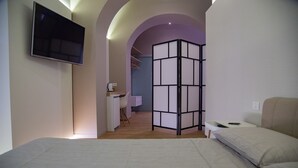 Et moderne hotelværelse med seng, skrivebord med stol, fjernsyn ophængt på væggen og en rumdeler med et garderobeskab bagved.