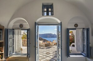 A room with an open door leading to a terrace with a table and chairs, a view of the sea, and a circular wall decoration. - Lava Oia's (Santorini)