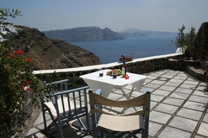 Una mesa con mantel blanco, una botella y un plato de comida en una terraza con pared de piedra y vistas al mar y las montañas.