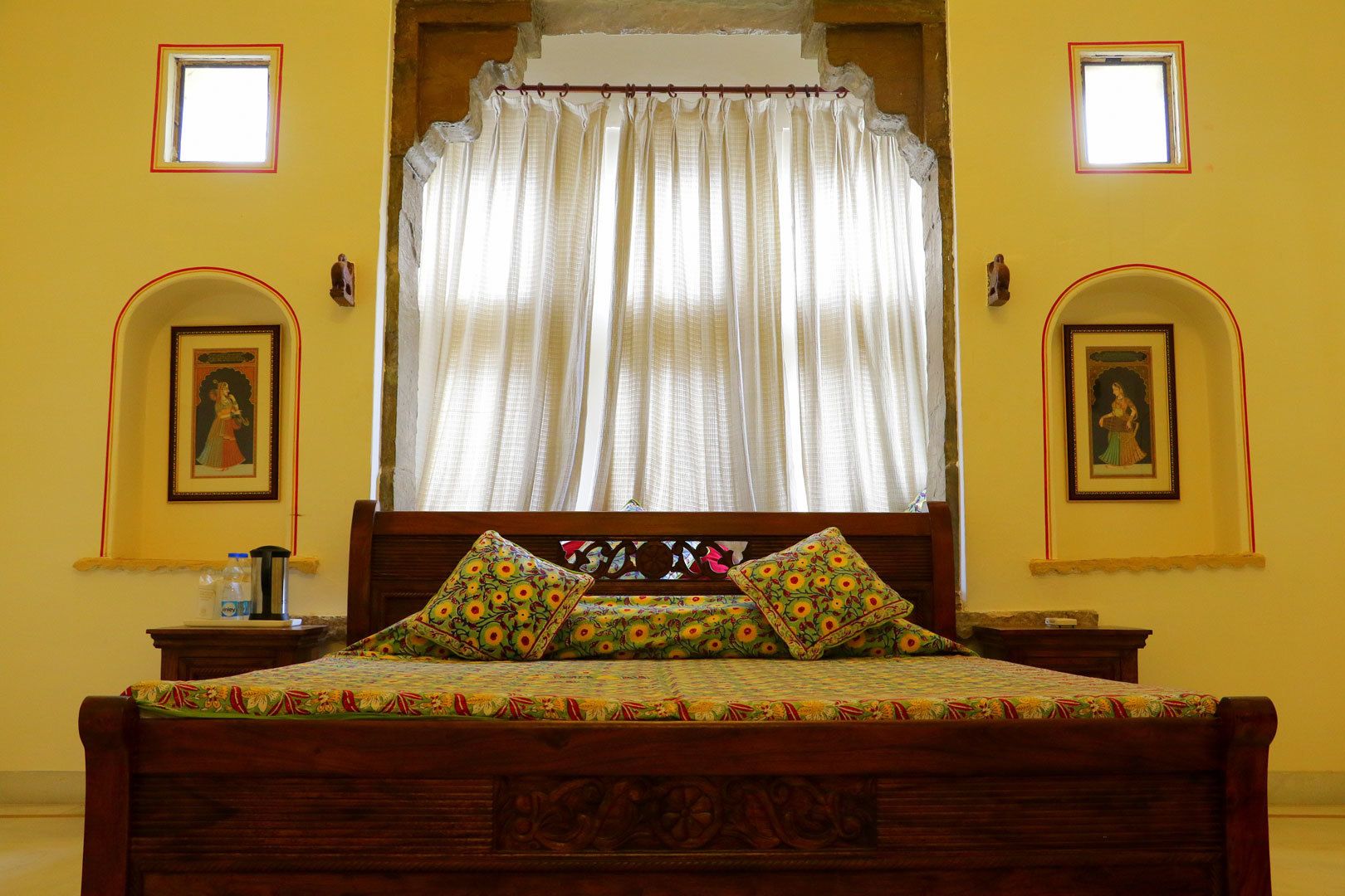 Un dormitorio tradicional con cama de madera, colcha estampada y cortinas. Hay dos marcos de cuadros en la pared y una mesita con una botella y un recipiente.