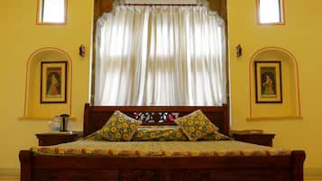 Un dormitorio tradicional con cama de madera, colcha estampada y cortinas. Hay dos marcos de cuadros en la pared y una mesita con una botella y un recipiente.