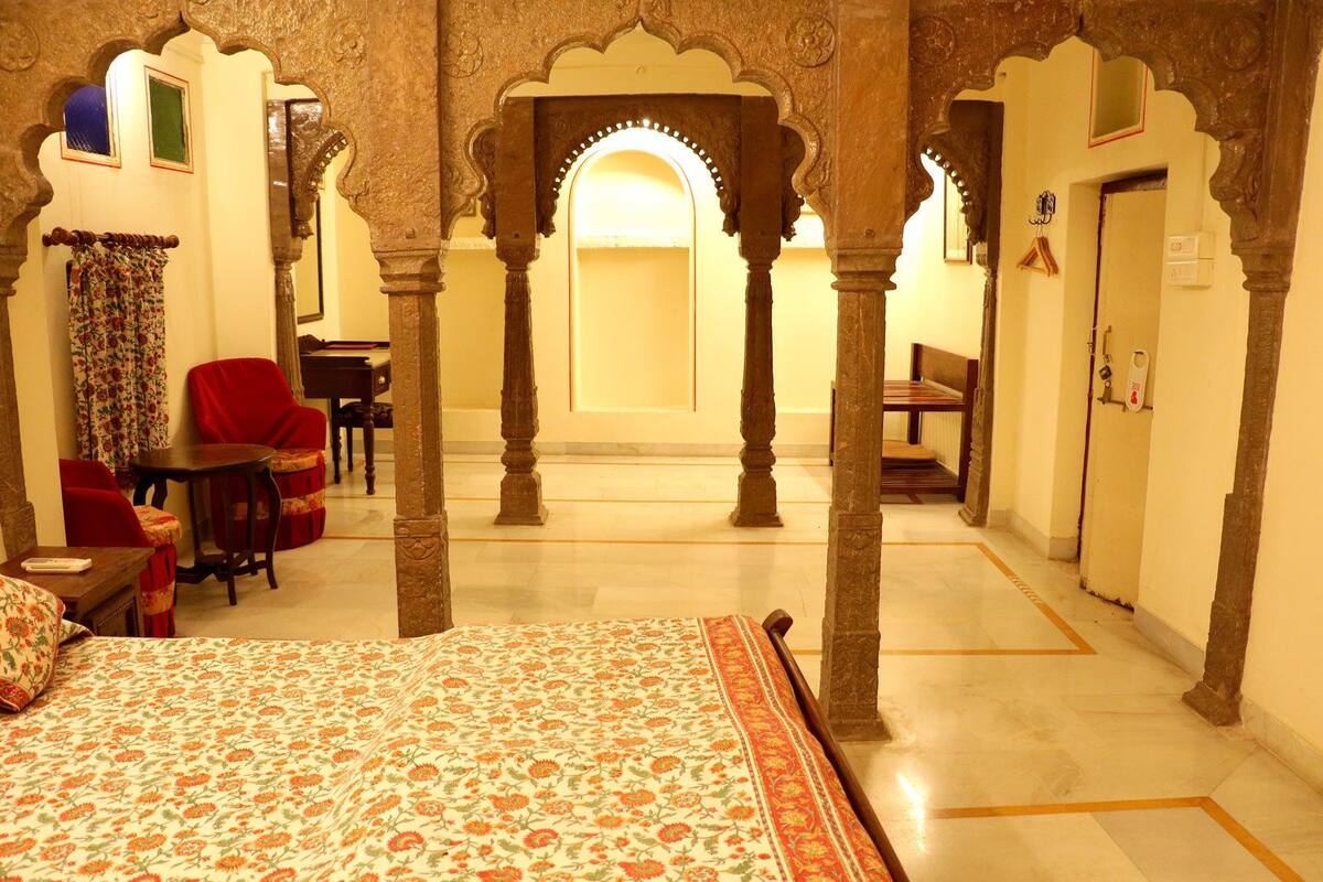 A room with a bed, red chairs, a table, and a small desk. There are ornate pillars and a doorway leading to another room.