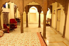 A room with a bed, red chairs, a table, and a small desk. There are ornate pillars and a doorway leading to another room.