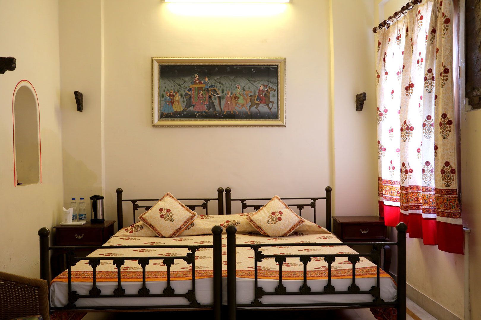 Una cama con una colcha estampada y dos almohadas, una mesita de noche con una botella de agua y un termo, un cuadro enmarcado en la pared y una ventana con cortinas florales.