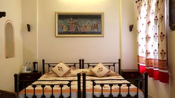 Una cama con una colcha estampada y dos almohadas, una mesita de noche con una botella de agua y un termo, un cuadro enmarcado en la pared y una ventana con cortinas florales.