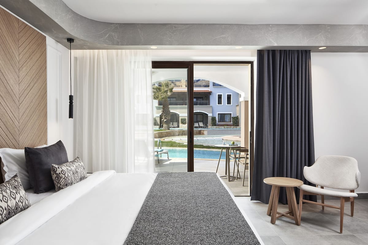 A modern hotel room with a large bed, a small table, a chair, and a view of a pool and buildings outside.