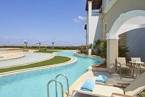 A pool area with a curved design, lounge chairs, and a table with towels. - Atlantica Caldera Palace - All Inclusive (Hersonissos)