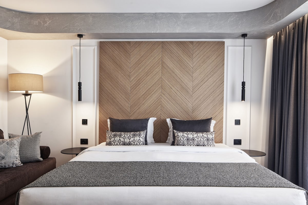 A modern hotel room with a large bed, a wooden headboard, two bedside tables, a floor lamp, and a view of the city.