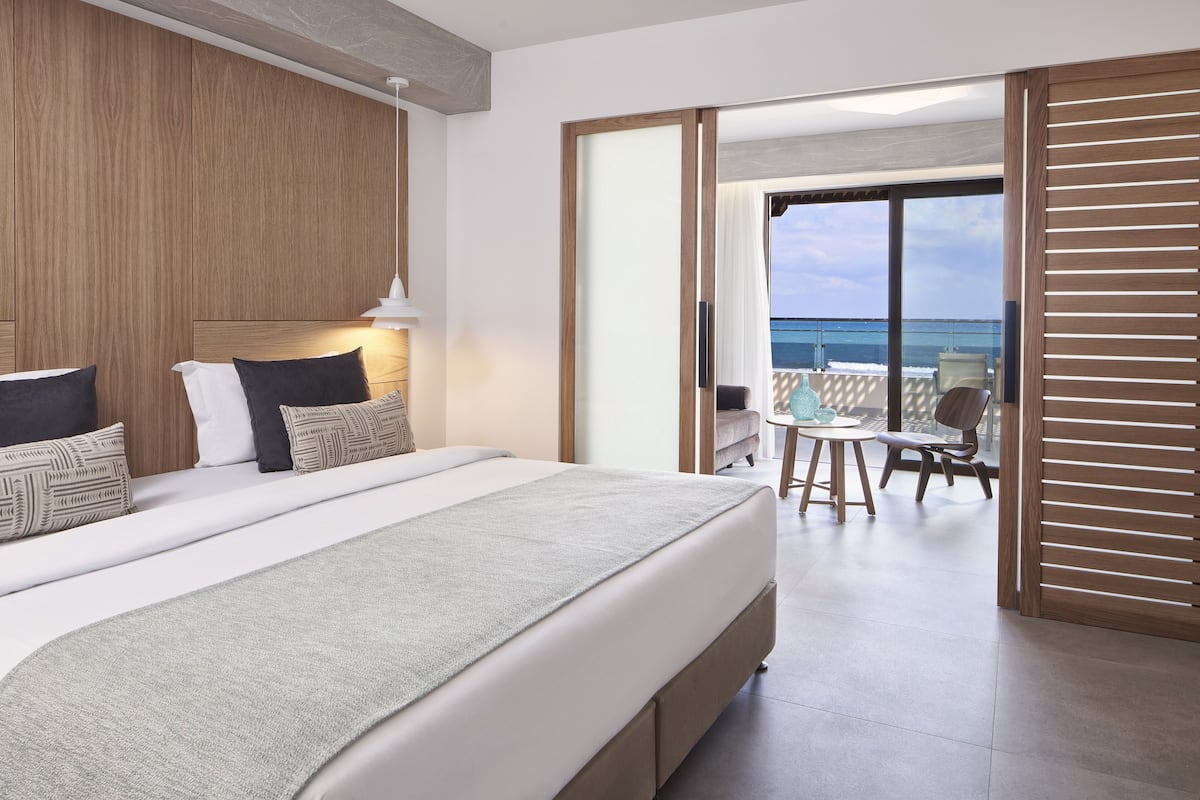 A modern hotel room with a large bed, a balcony with a view of the sea, and a sliding door leading to an outdoor seating area.