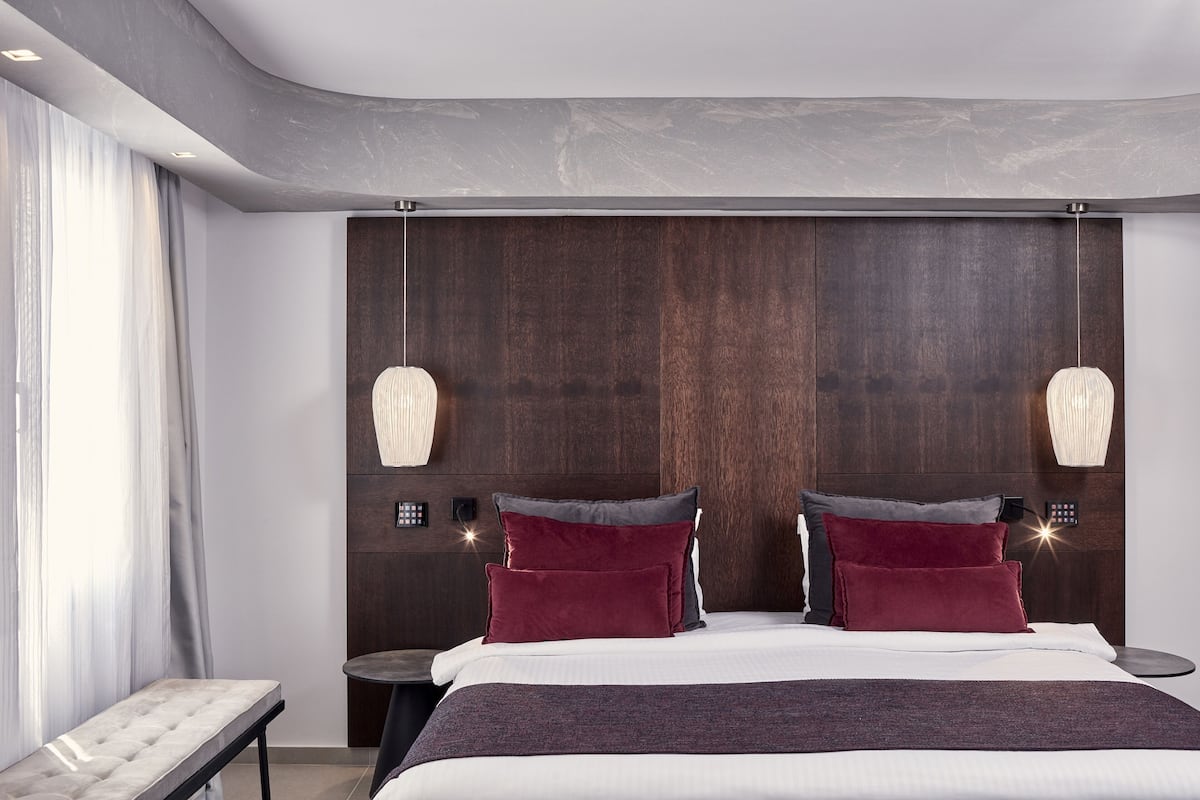 A modern hotel room with a large bed, dark wood paneling, two bedside lamps, and a bench by the window.