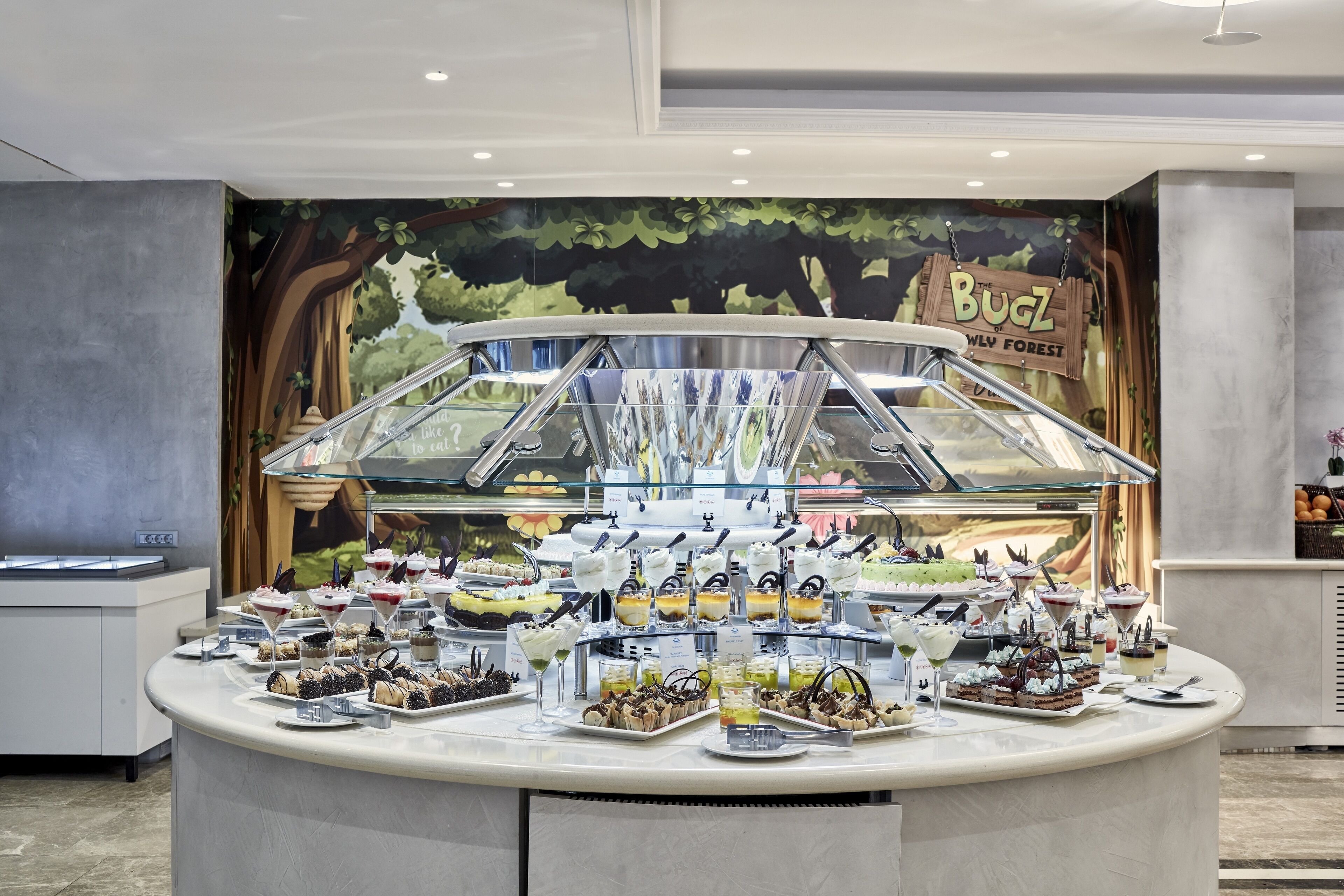 A buffet with a variety of desserts and drinks, a glass display case, and a mural in the background.