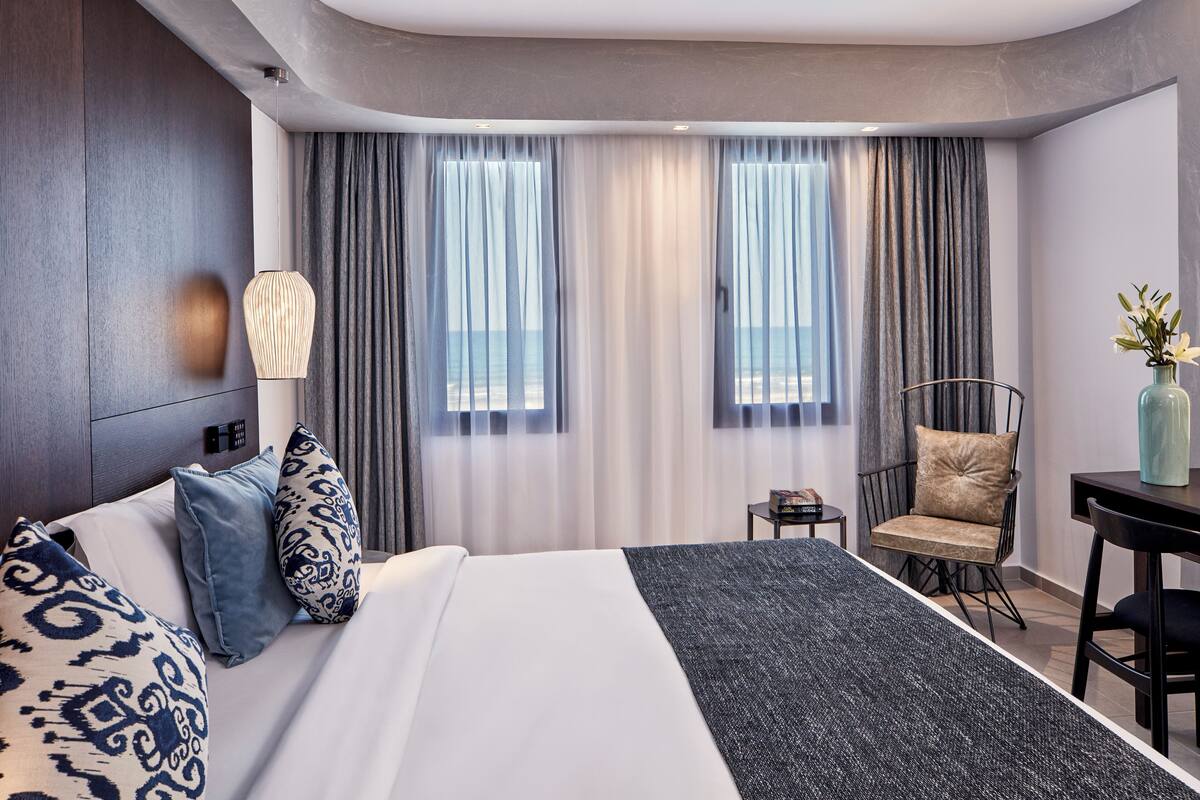A hotel room with a large bed, two windows with curtains, a small table with a vase, and a chair.