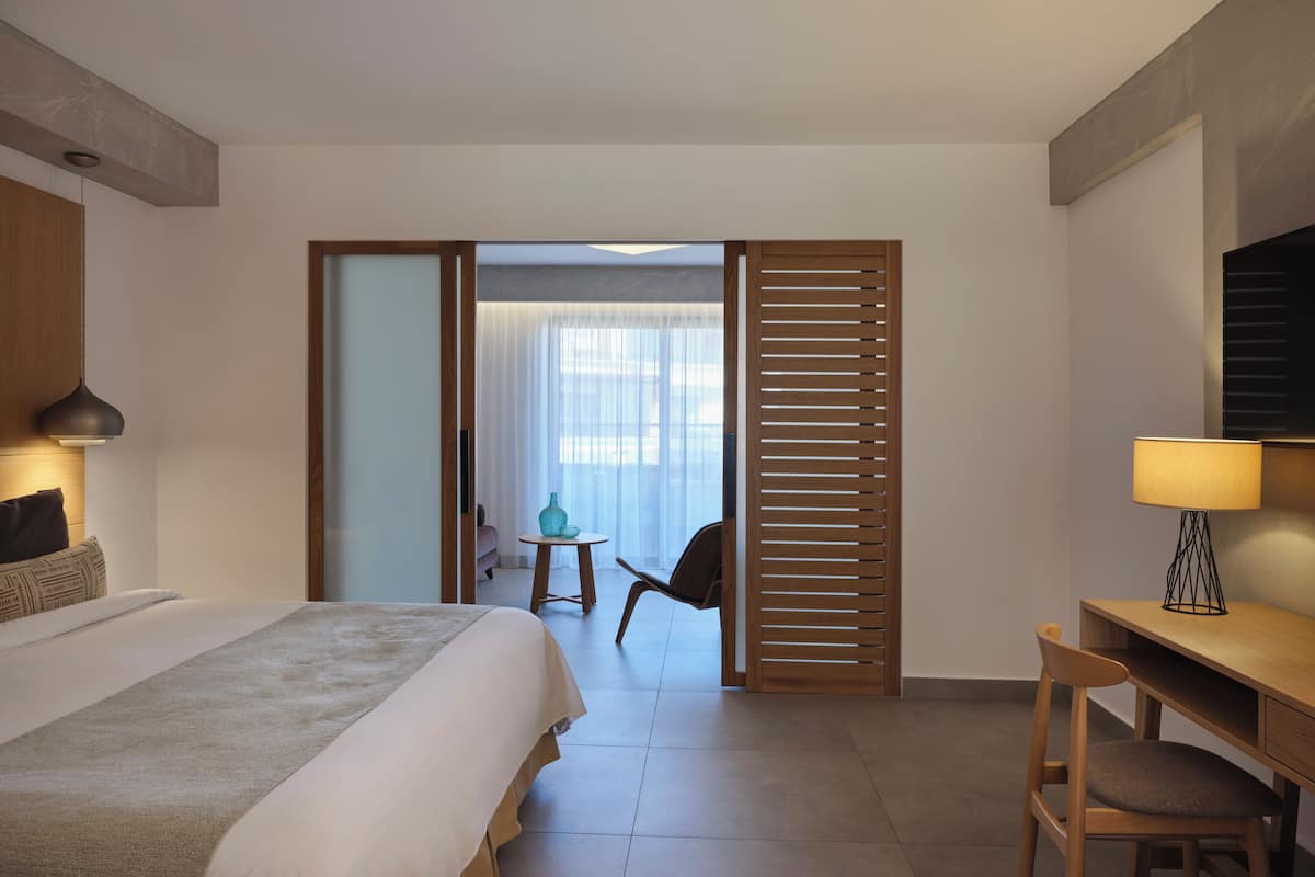 A hotel room with a bed, a desk, a chair, a lamp, and a view through a sliding door to a balcony.