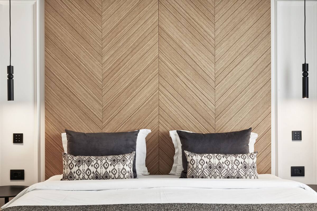 A hotel room with a herringbone-patterned wooden headboard, two patterned pillows, a white bedspread, and black pendant lights.