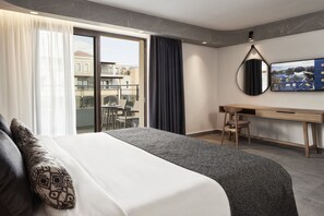 A modern hotel room with a large bed, a desk, and a balcony with a view of buildings. - Atlantica Caldera Palace - All Inclusive (Hersonissos)