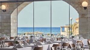 Restaurante com mesas preparadas para refeições, grandes janelas e vista para o mar.