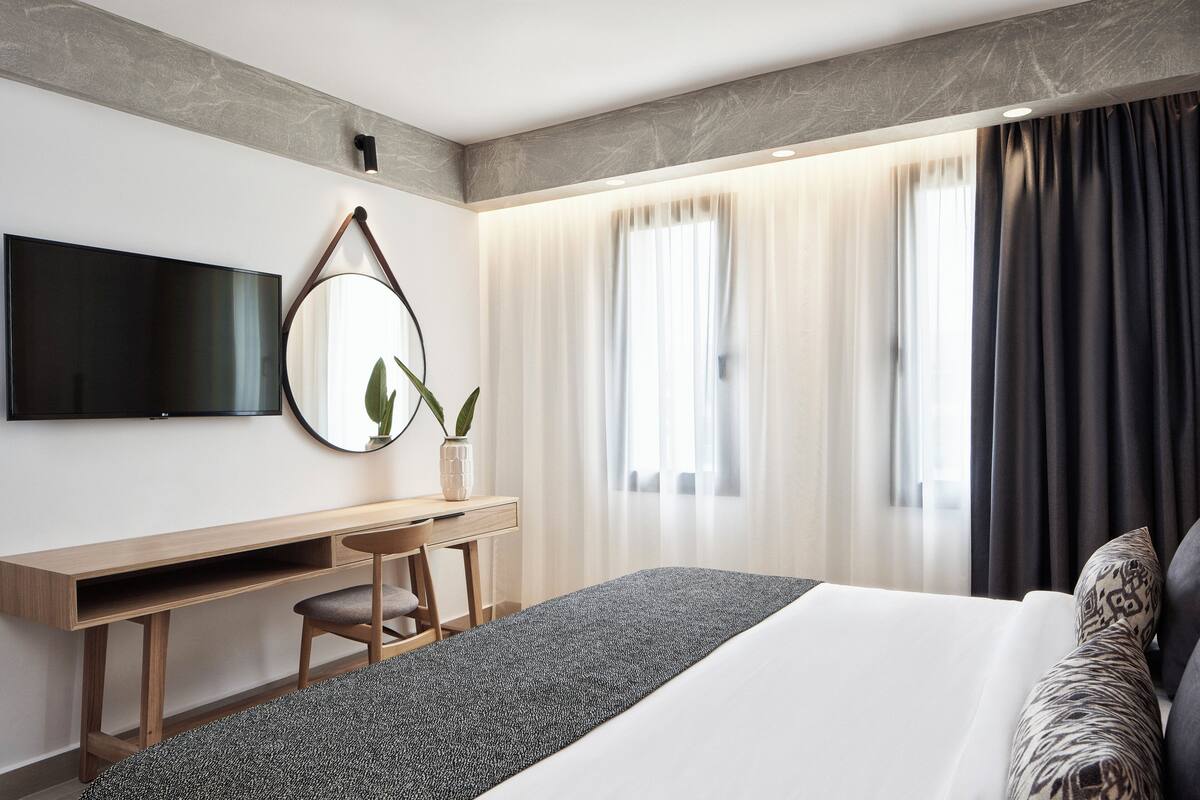 A modern hotel room with a bed, a desk, a chair, a TV, and a large window with curtains.