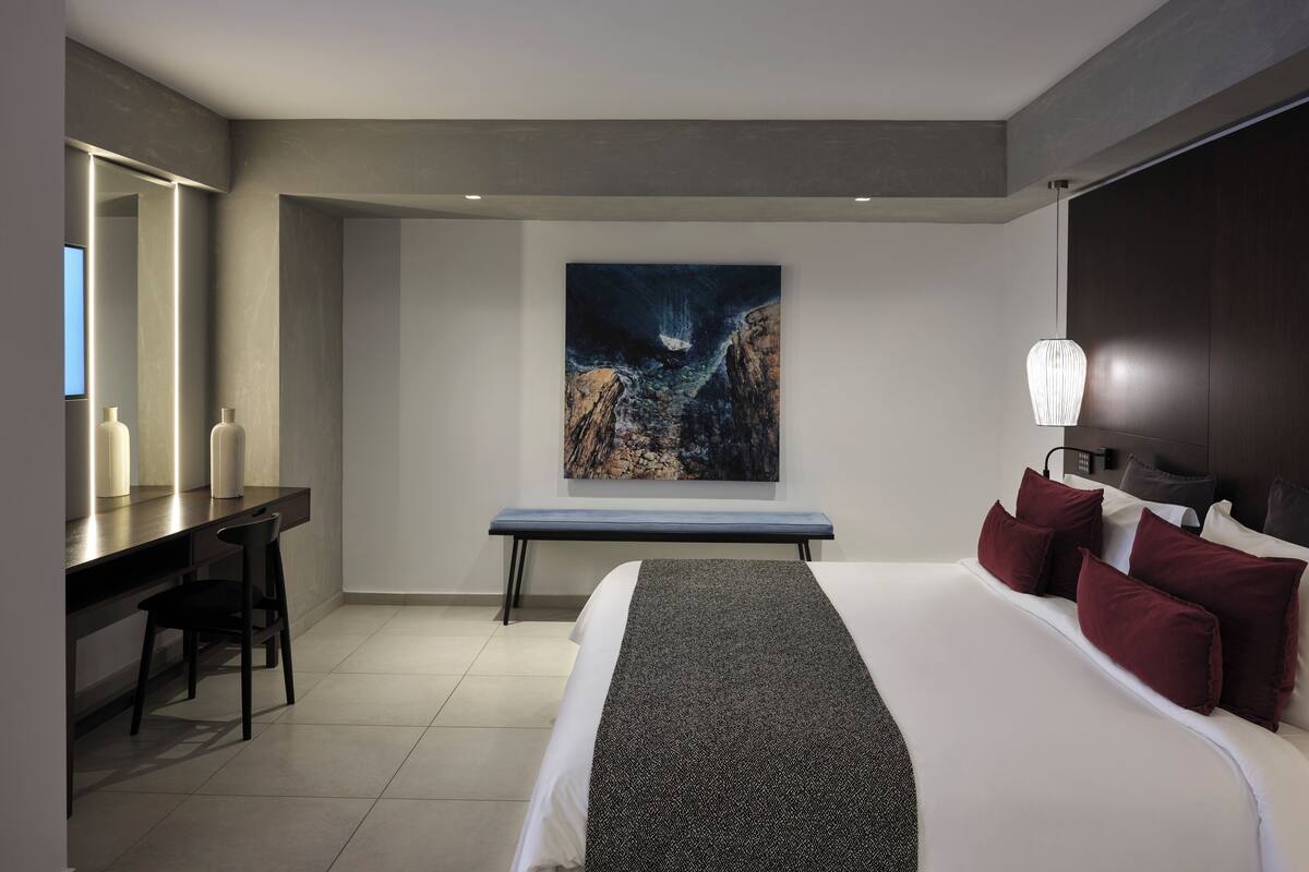 A modern hotel room with a large bed, a desk, and a painting on the wall.