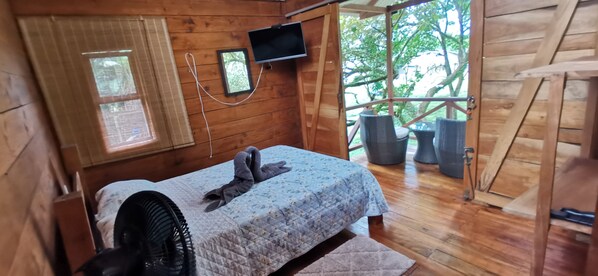 Een houten slaapkamer met een bed, een aan de muur bevestigde televisie en een raam met uitzicht op groen.