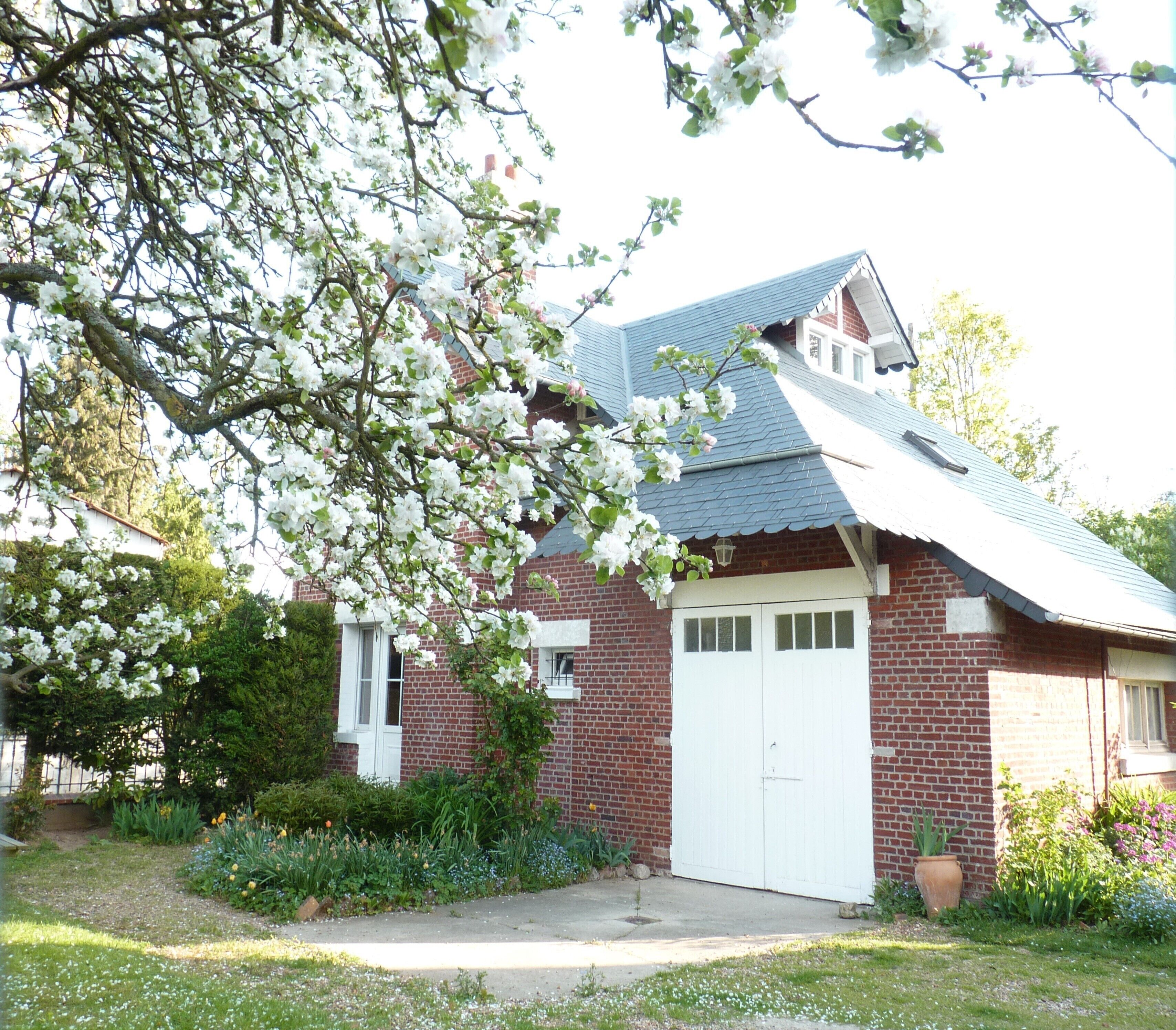 Une maison en briques avec une porte de garage blanche, entourée d’un jardin où poussent différentes plantes et fleurs.