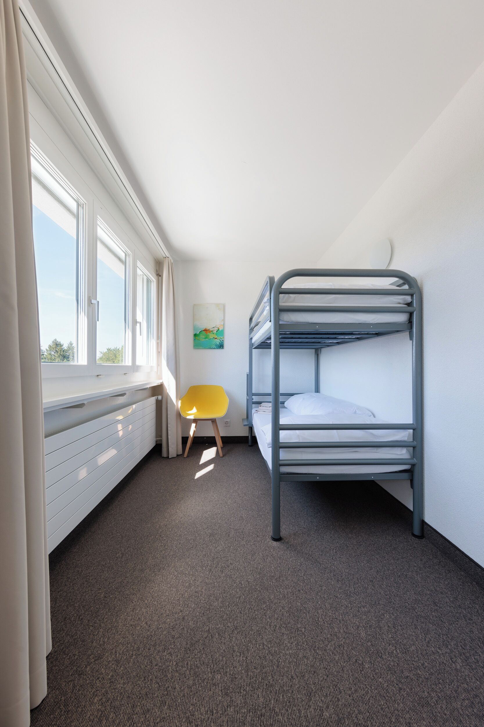 Ein Schlafzimmer mit Etagenbetten, einem Heizkörper, einem Fenster mit Vorhängen, einem gelben Stuhl und einem Bild an der Wand.