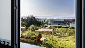 A view from a window of a garden area with a wooden pergola, a table, and chairs, and a backdrop of buildings and mountains. - Domaine NDR (Villars-sur-Glane)