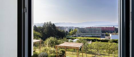 Vista desde una ventana a un jardín con un porche de madera, una mesa y sillas, y edificios y montañas al fondo.