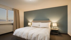 A neatly made bed with white linens, two bedside tables with lamps, a window with curtains, and a view of the mountains. - Domaine NDR (Villars-sur-Glane)