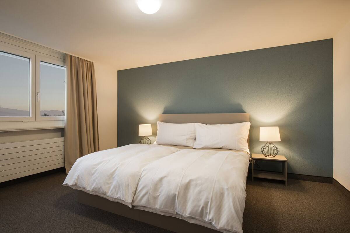 A neatly made bed with white linens, two bedside tables with lamps, a window with curtains, and a view of the mountains.