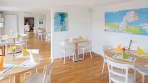 A modern dining area with white chairs and tables, featuring abstract art on the walls and a wooden floor. - Domaine NDR (Villars-sur-Glane)