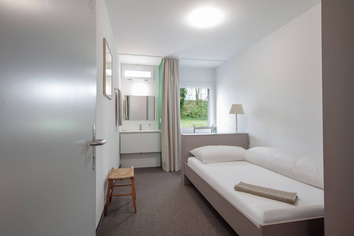 A hotel room with a bed, a small table, a chair, a window with a view of greenery, and a bathroom with a sink and mirror.