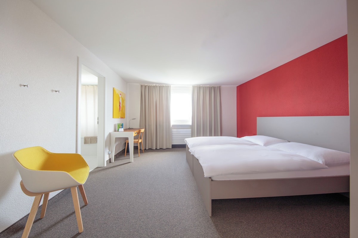 A hotel room with a bed, a desk, a chair, and a window with curtains.