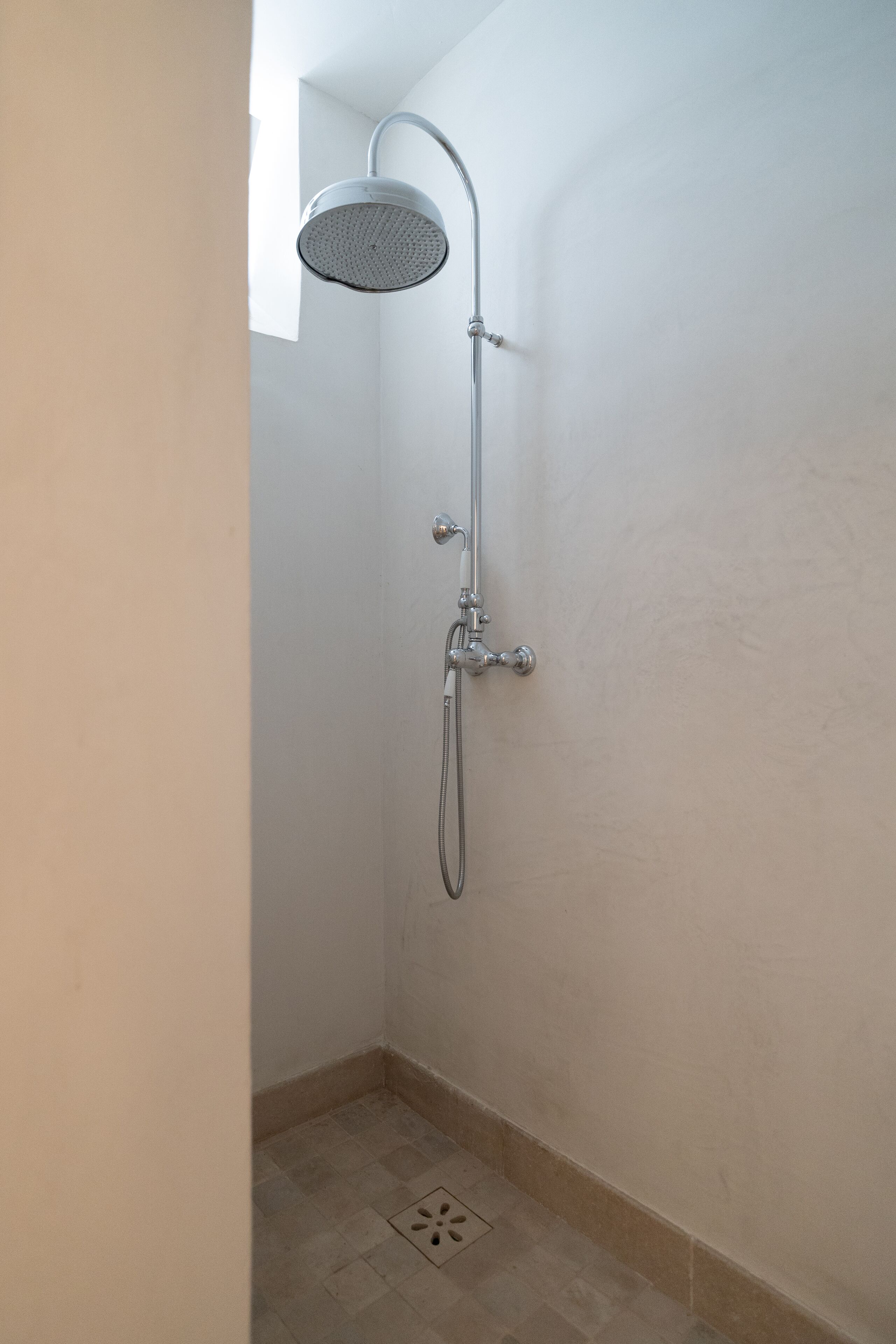 A shower with a handheld showerhead and a wall-mounted faucet in a corner of a room with plain walls and a tiled floor.