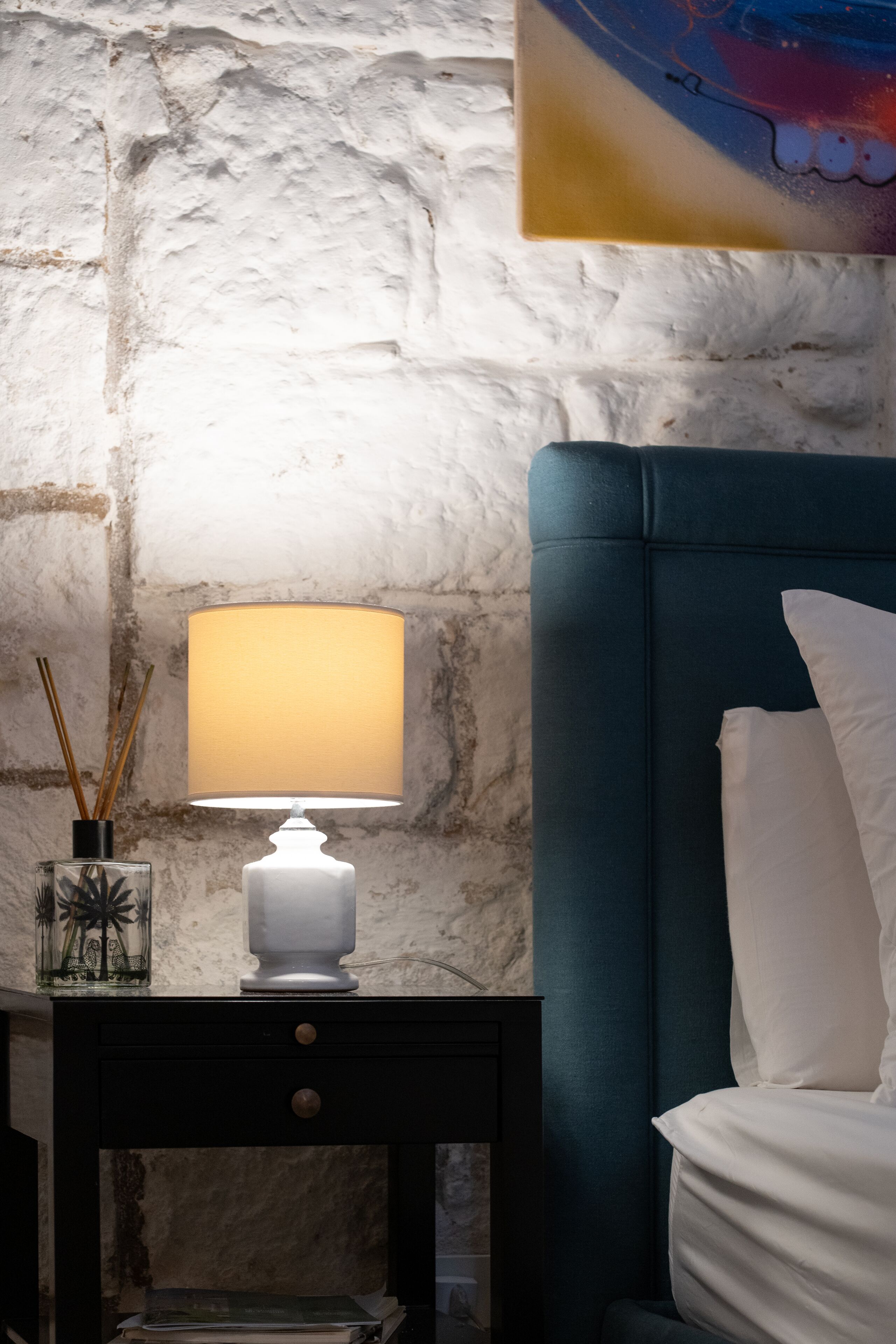 Ein Schlafzimmer mit einem türkisblauen Kopfteil, weißer Bettwäsche, einem Nachttisch mit Lampe und einer Wand mit strukturierter Steinoptik.
