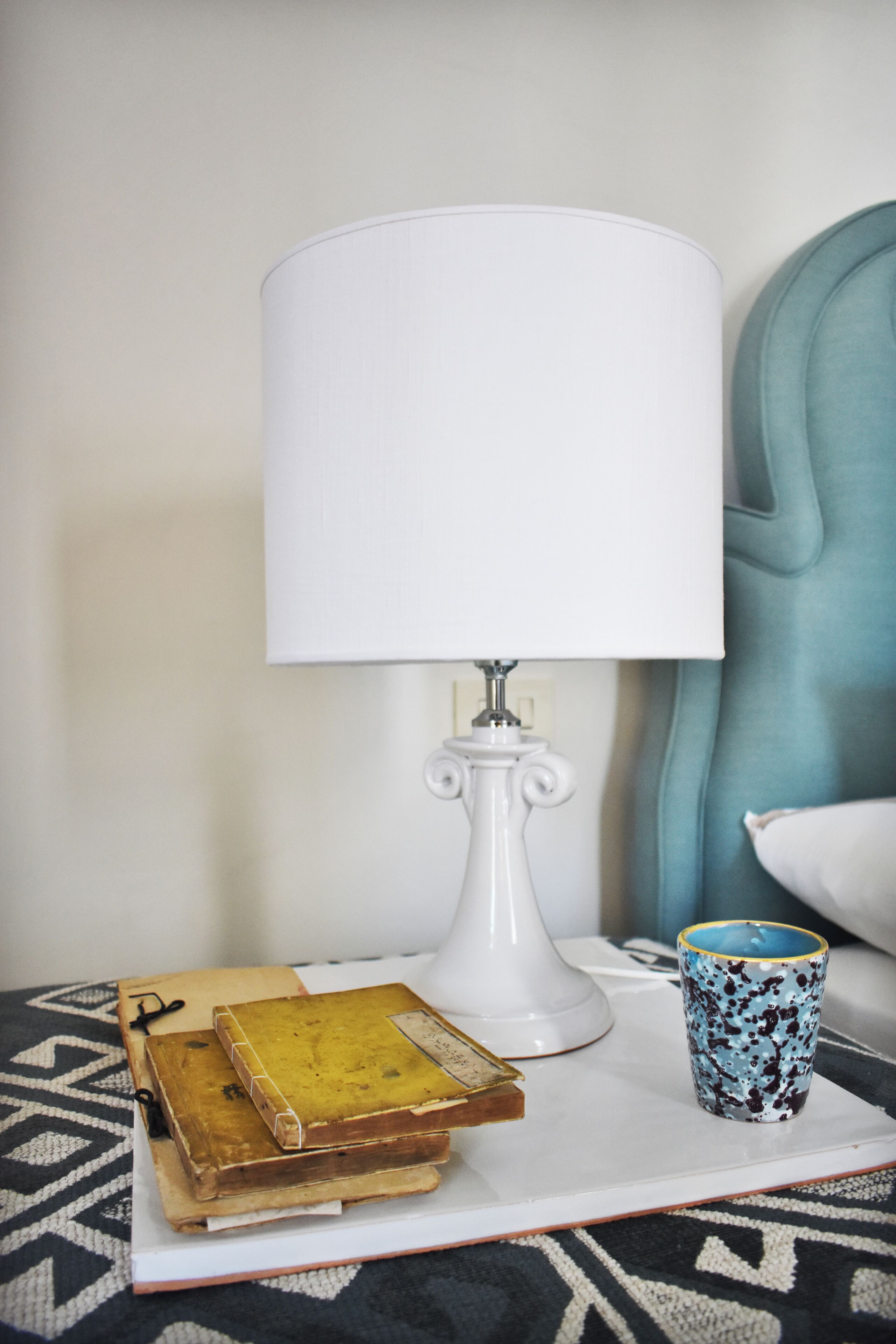 A bedside table with a white lamp, a blue mug, and several books.